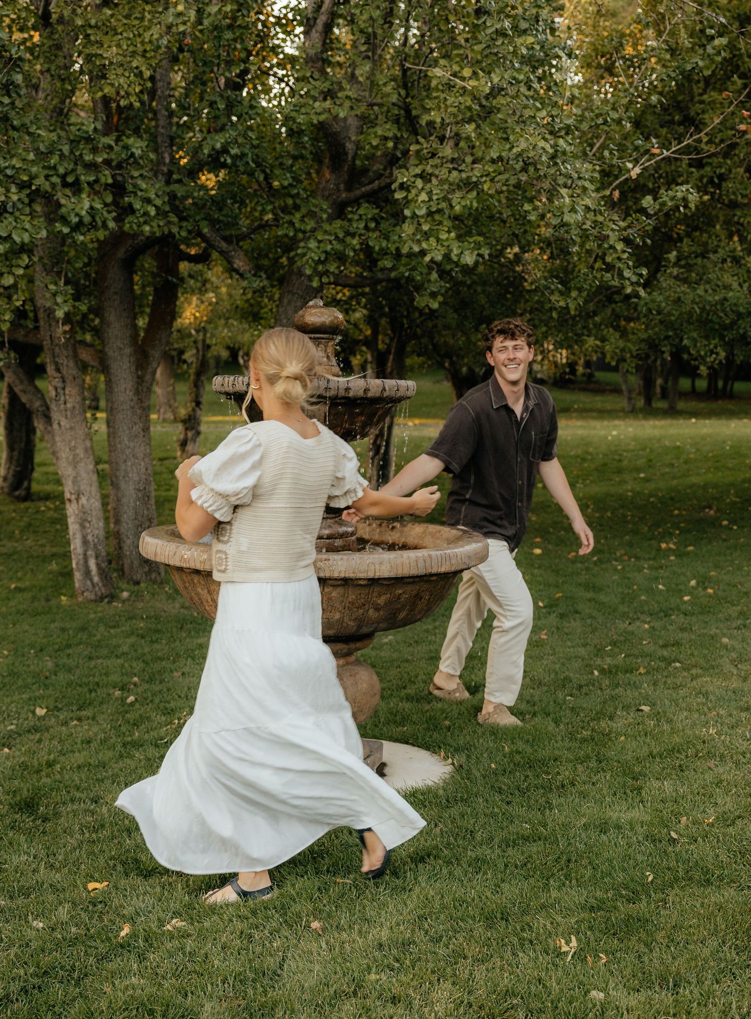 Couple playfully chasing each other around a rustic fountain in a lush green park during autumn.