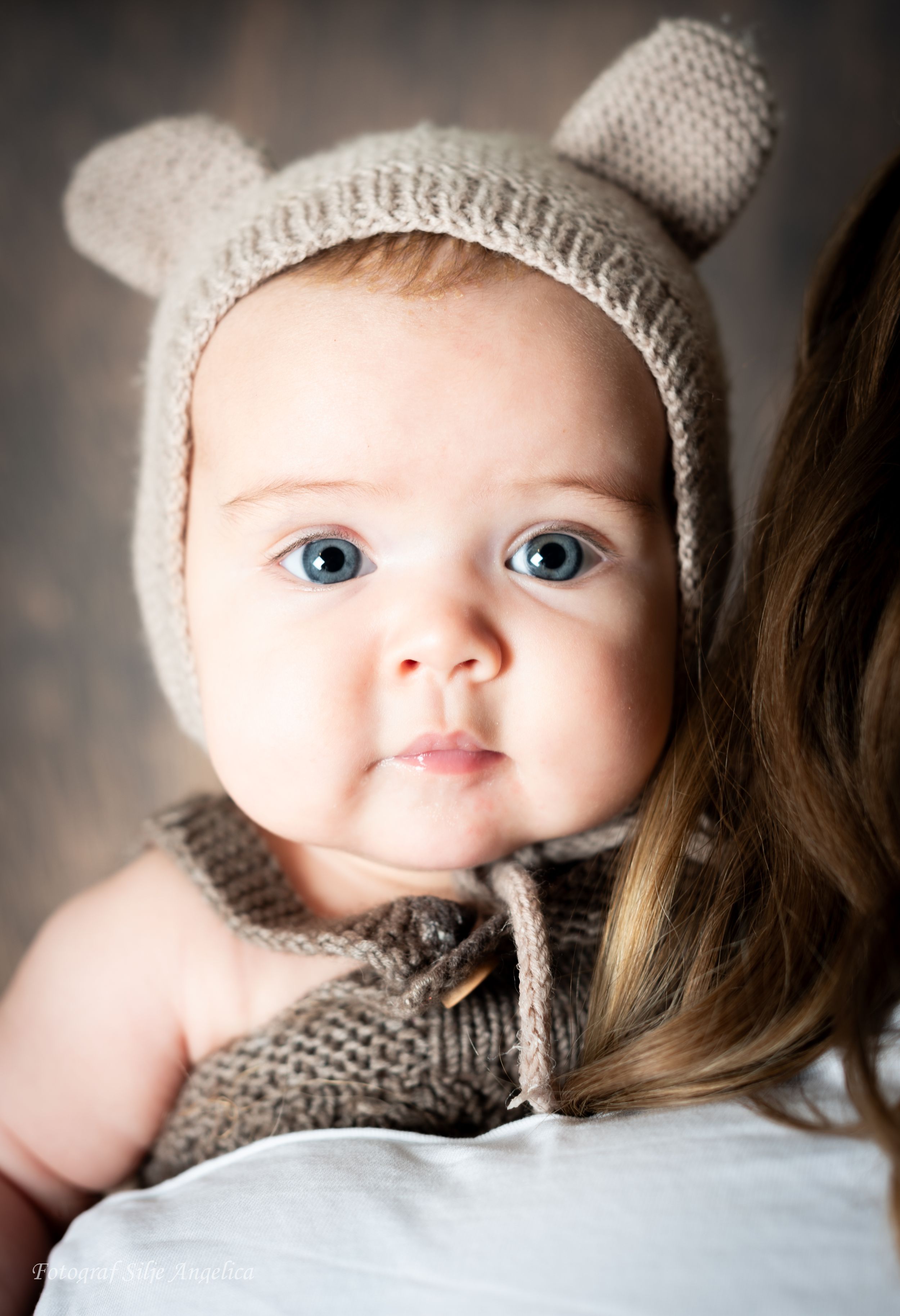 Baby wearing knitted bear hat and sweater looking at camera with bright blue eyes.