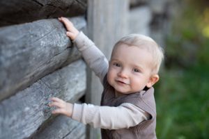 Small child in neutral colored sweater reaching up wooden wall outdoors.