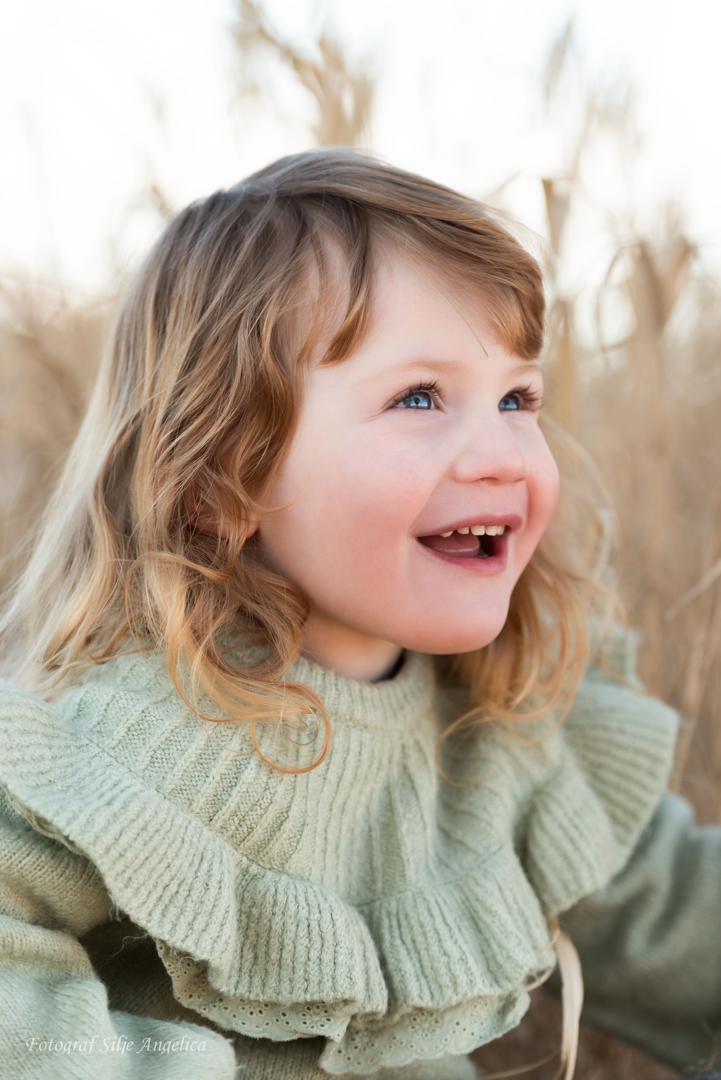 Smiling toddler wearing sage green ruffled sweater against blurred natural background.