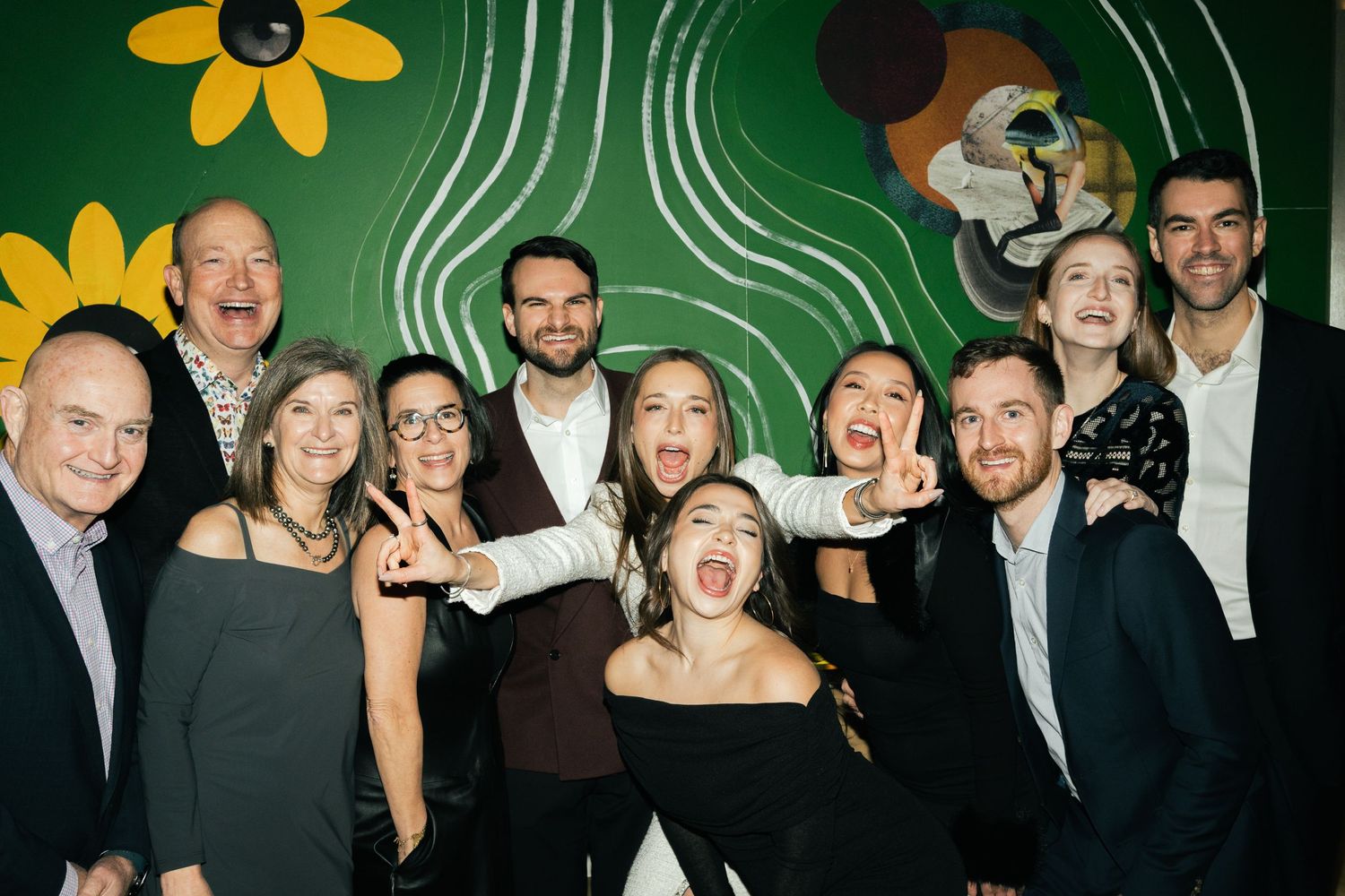 A lively group photo with people posing playfully against a green wall decorated with yellow sunflower designs.