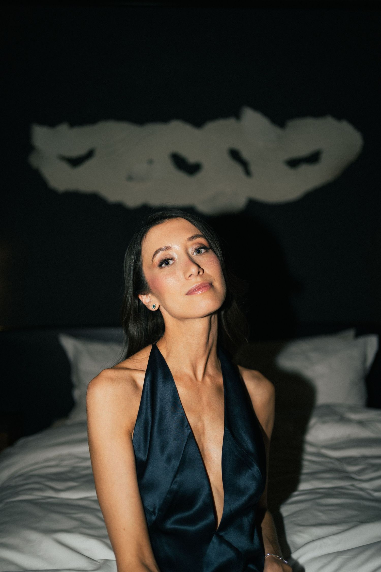 Woman in navy blue dress seated on white bedding with moody lighting.