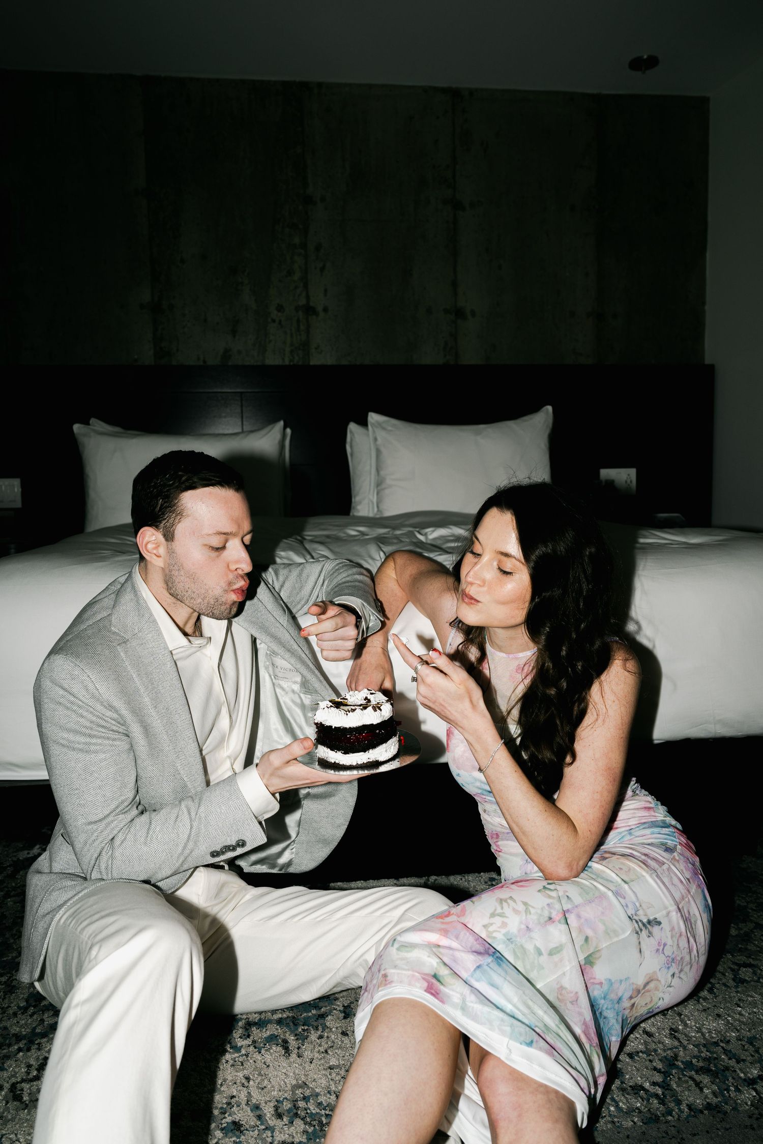 Couple sharing a small cake while sitting together on a hotel bed.
