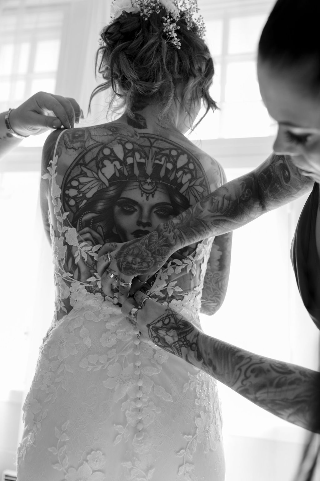 Black and white photo of a bride getting dressed, showing an intricate back tattoo through delicate wedding dress lace.