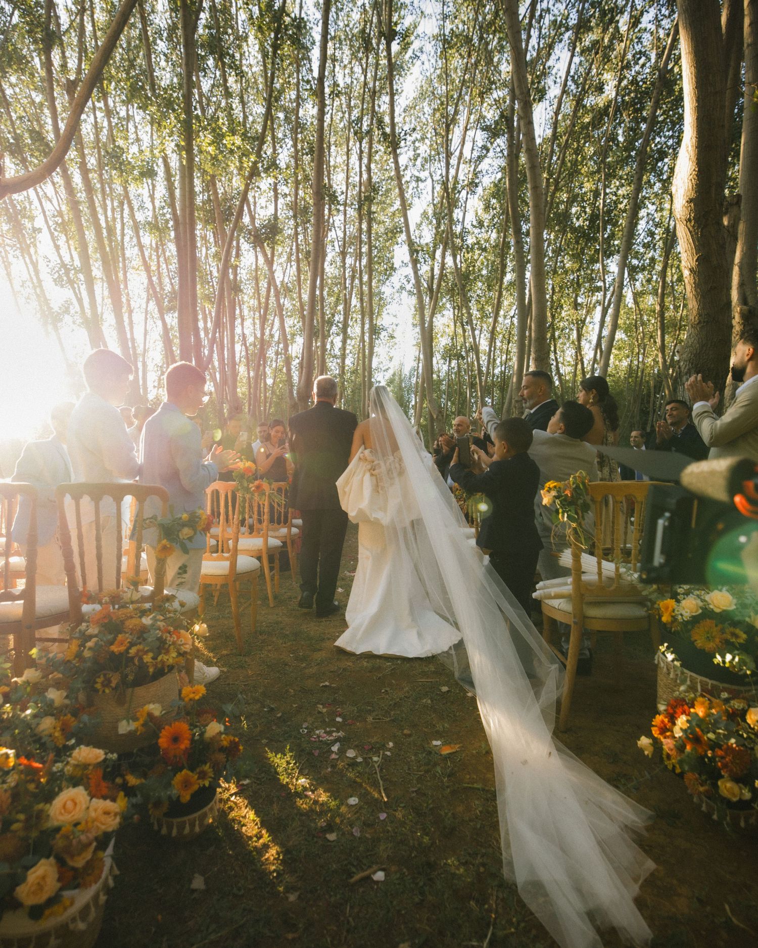 Outdoor forest wedding ceremony with sunlight streaming through trees and orange floral decorations.