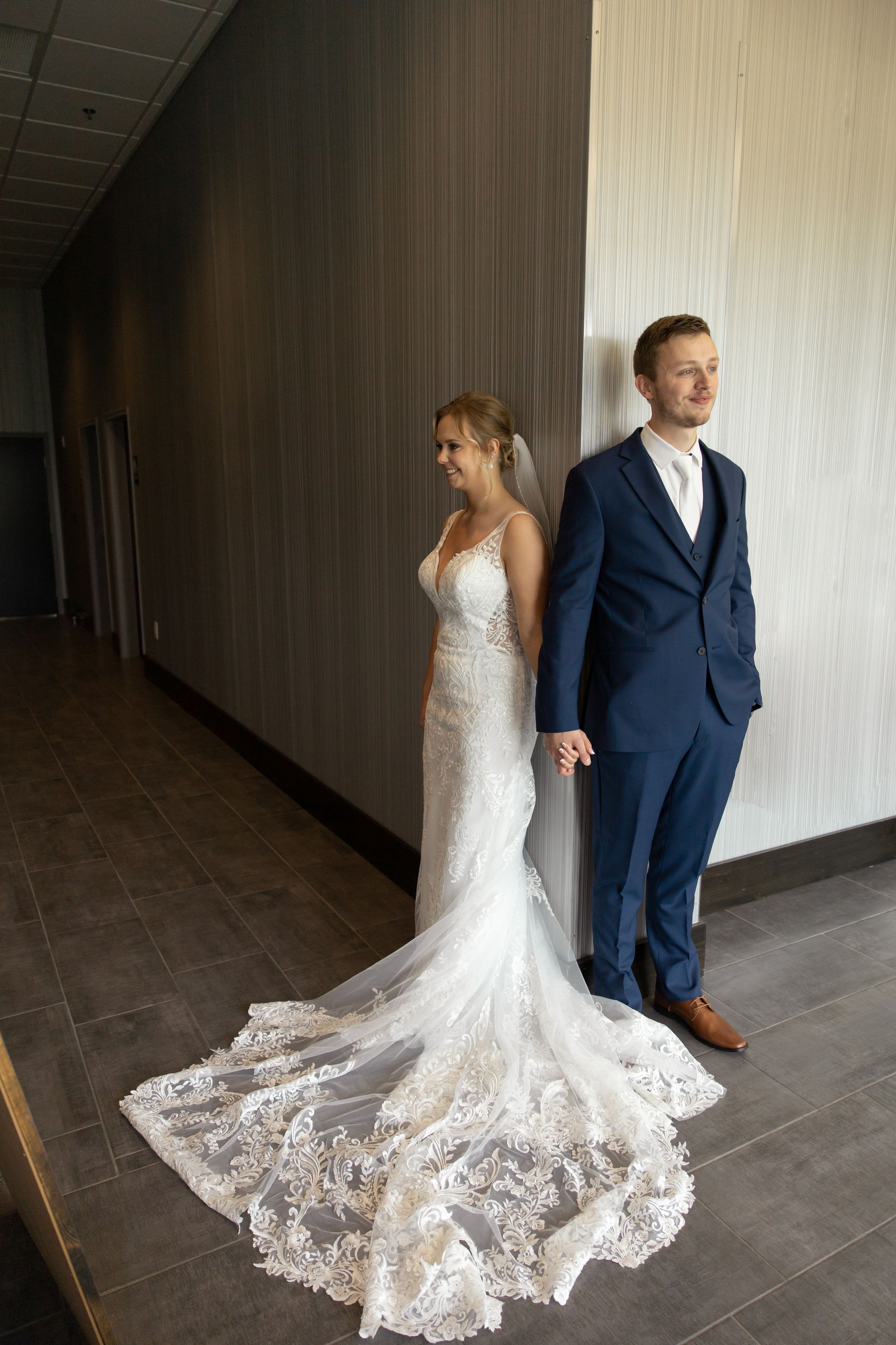 Sioux City, Iowa photographer. Bride in lace wedding gown with train stands with groom in navy suit against modern wall.