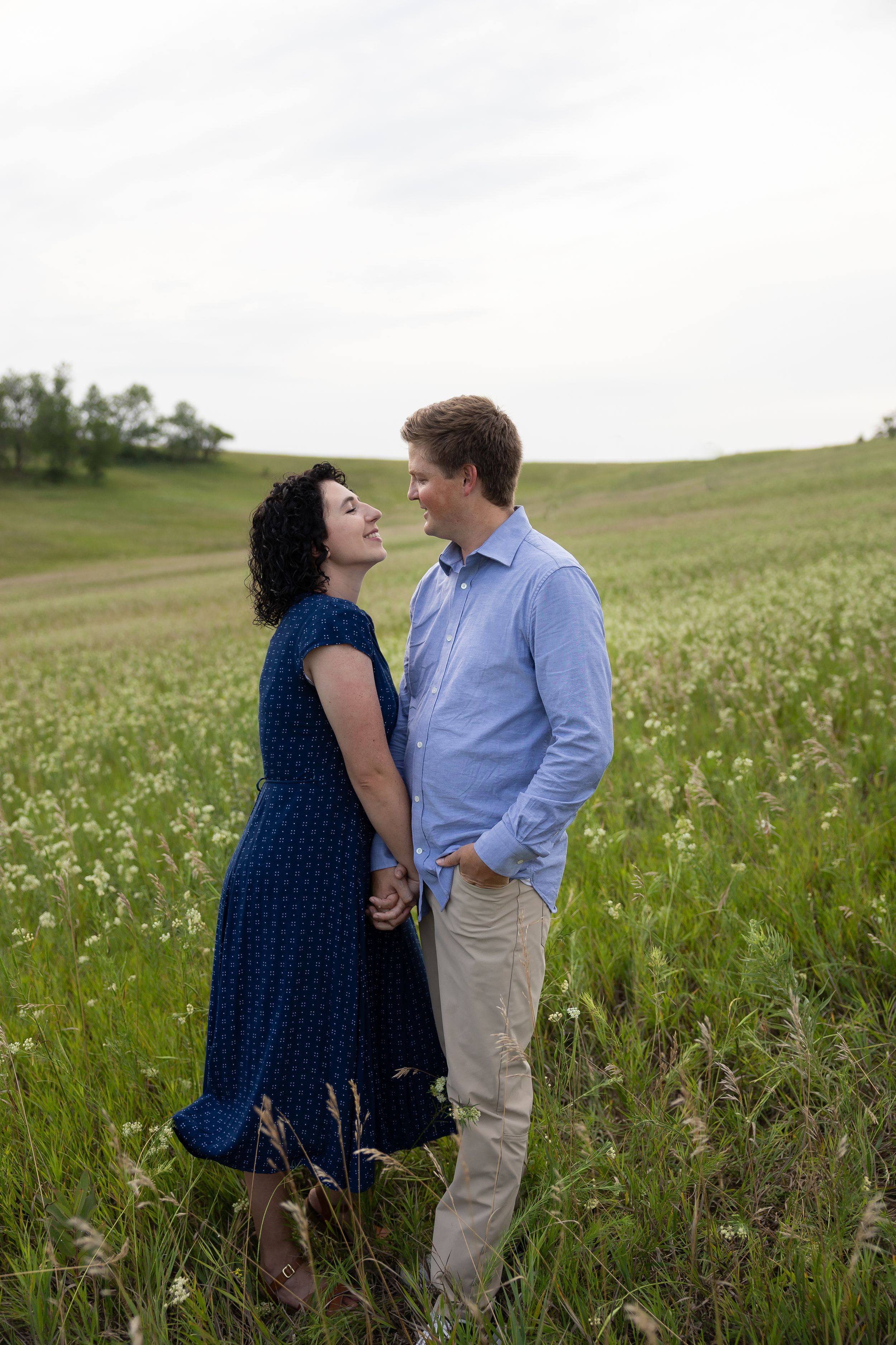 Sioux City, Iowa photographer. Couple holding hands and smiling at each other in a lush green field with wildflowers on a cloudy day.