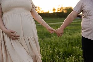Sioux City, Iowa photographer. A pregnant woman in a beige dress holds hands with her partner in a lush green field at sunset.