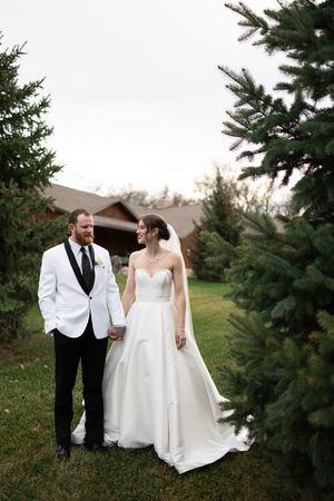 Sioux City, Iowa photographer. Bride and groom hold hands and share a smile while standing among tall evergreen trees outdoors.
