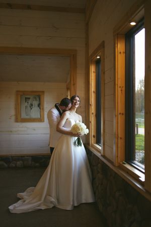 Sioux City, Iowa photographer. Groom kisses bride on the cheek as she smiles holding a white bouquet inside a charming log cabin venue.