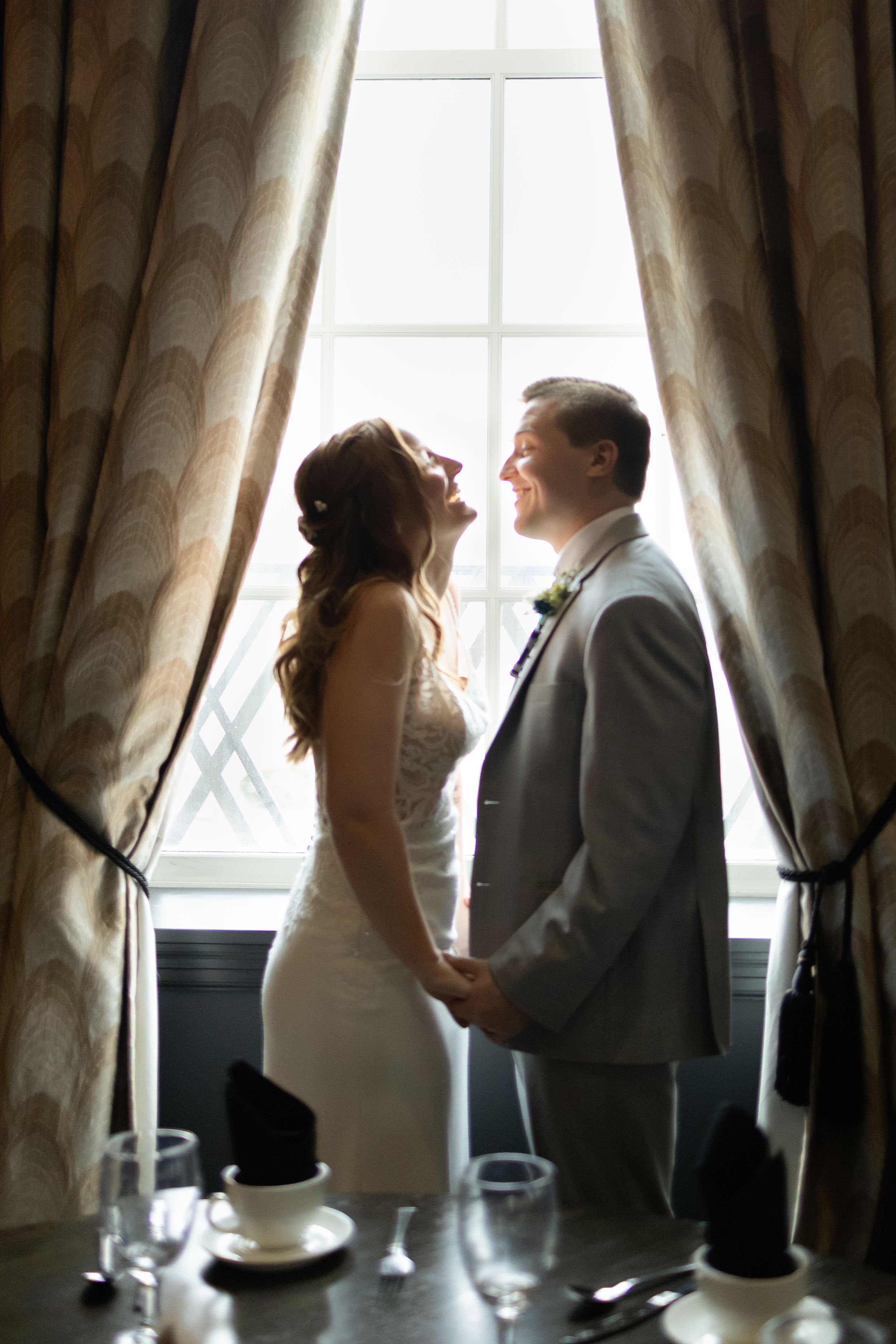 Sioux City, Iowa photographer. Bride and groom hold hands and smile at each other in silhouette before a tall window with draped curtains.