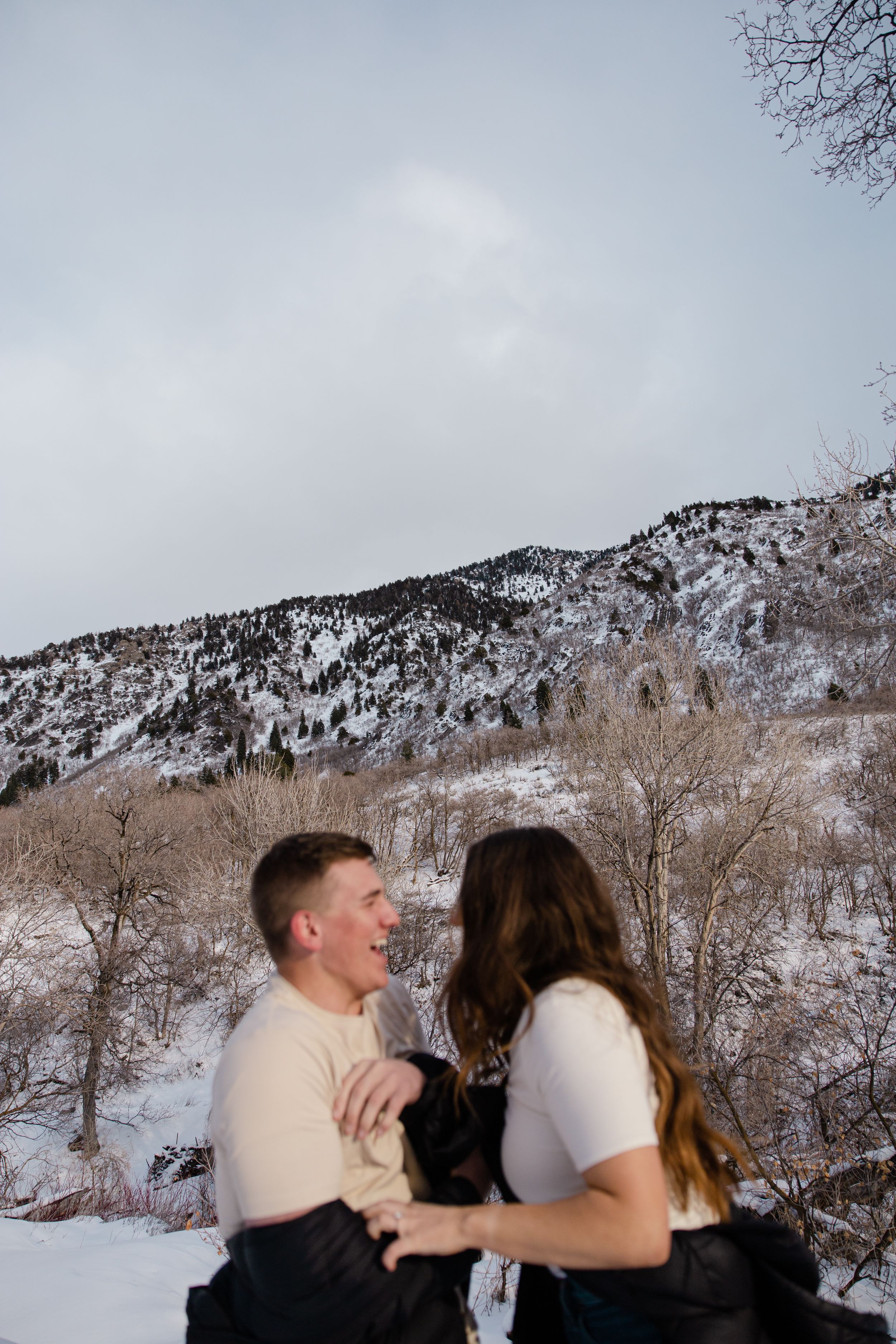 Sioux City, Iowa photographer. A smiling couple embraces outdoors in a snowy mountain landscape with bare winter trees behind them.