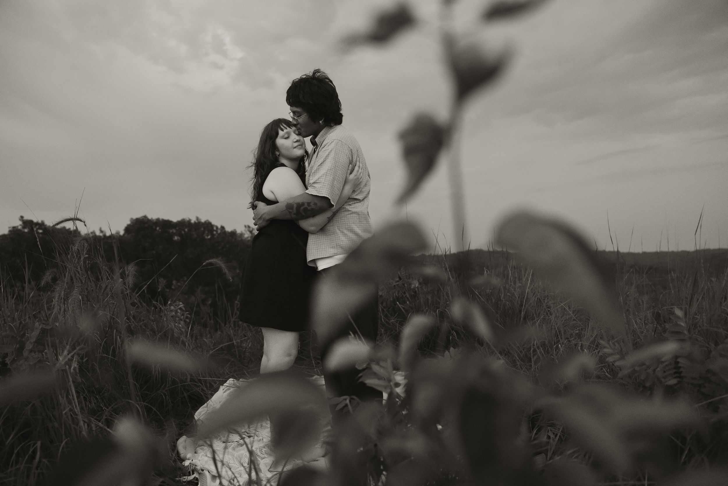 Black and white photo of a romantic couple embracing in a field, sharing a tender moment under a cloudy sky.