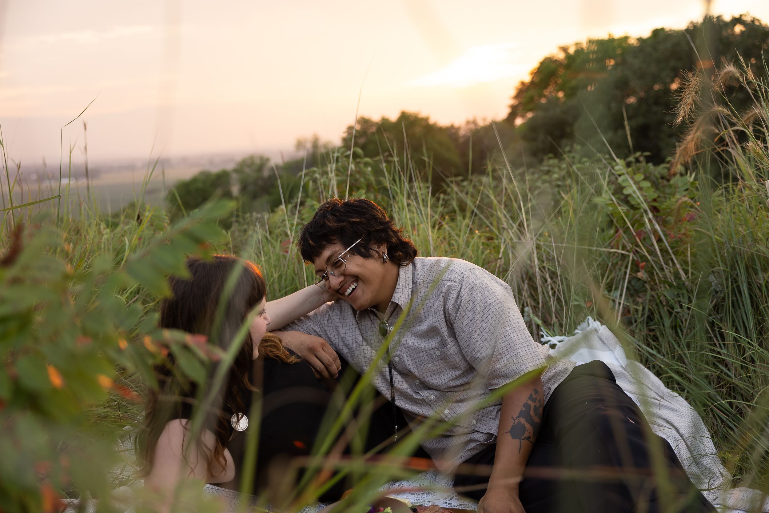 Couple sitting together in tall green grass at sunset, sharing a romantic outdoor picnic moment in nature.