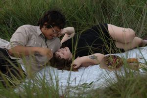 Couple lying together in tall green grass on a white blanket in a romantic outdoor setting.