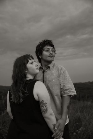 Black and white photo of a couple holding hands in a field, woman with arm tattoo leans on man wearing glasses.