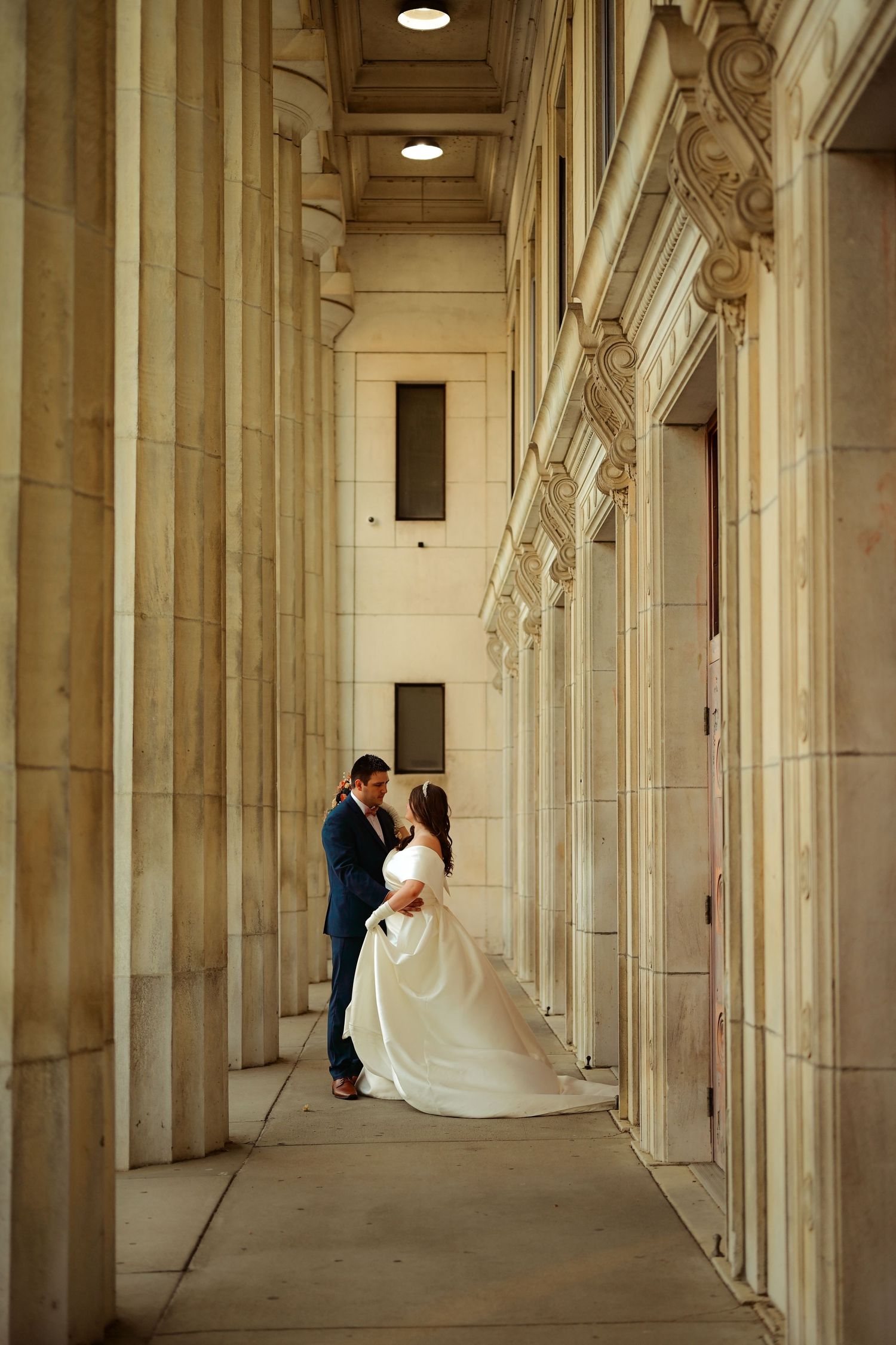 Elegant wedding couple stands in grand colonnade hallway with dramatic architectural columns and lighting.