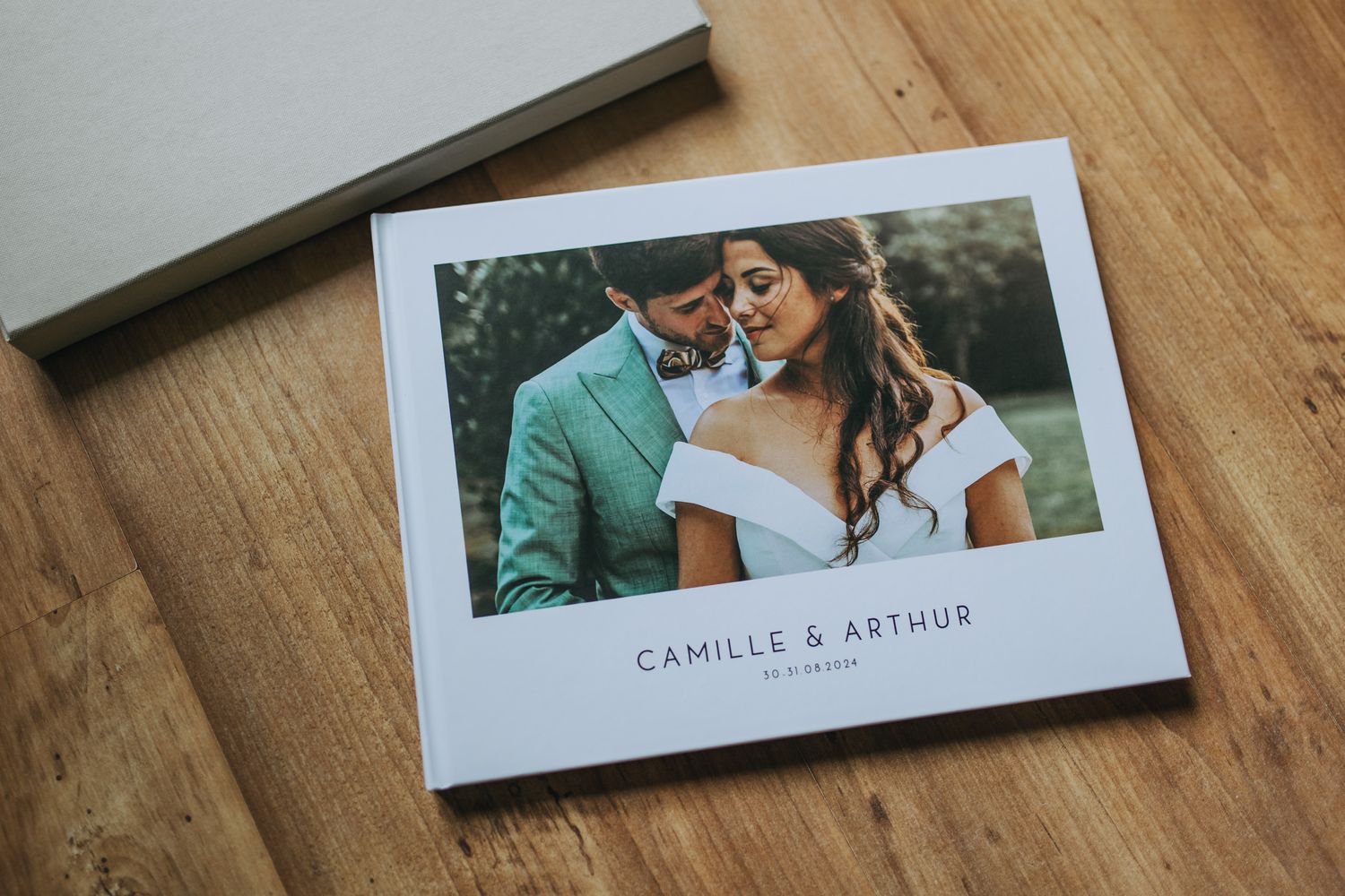 A wedding photo album with white cover lies on wooden surface showing couple in green and white attire.
