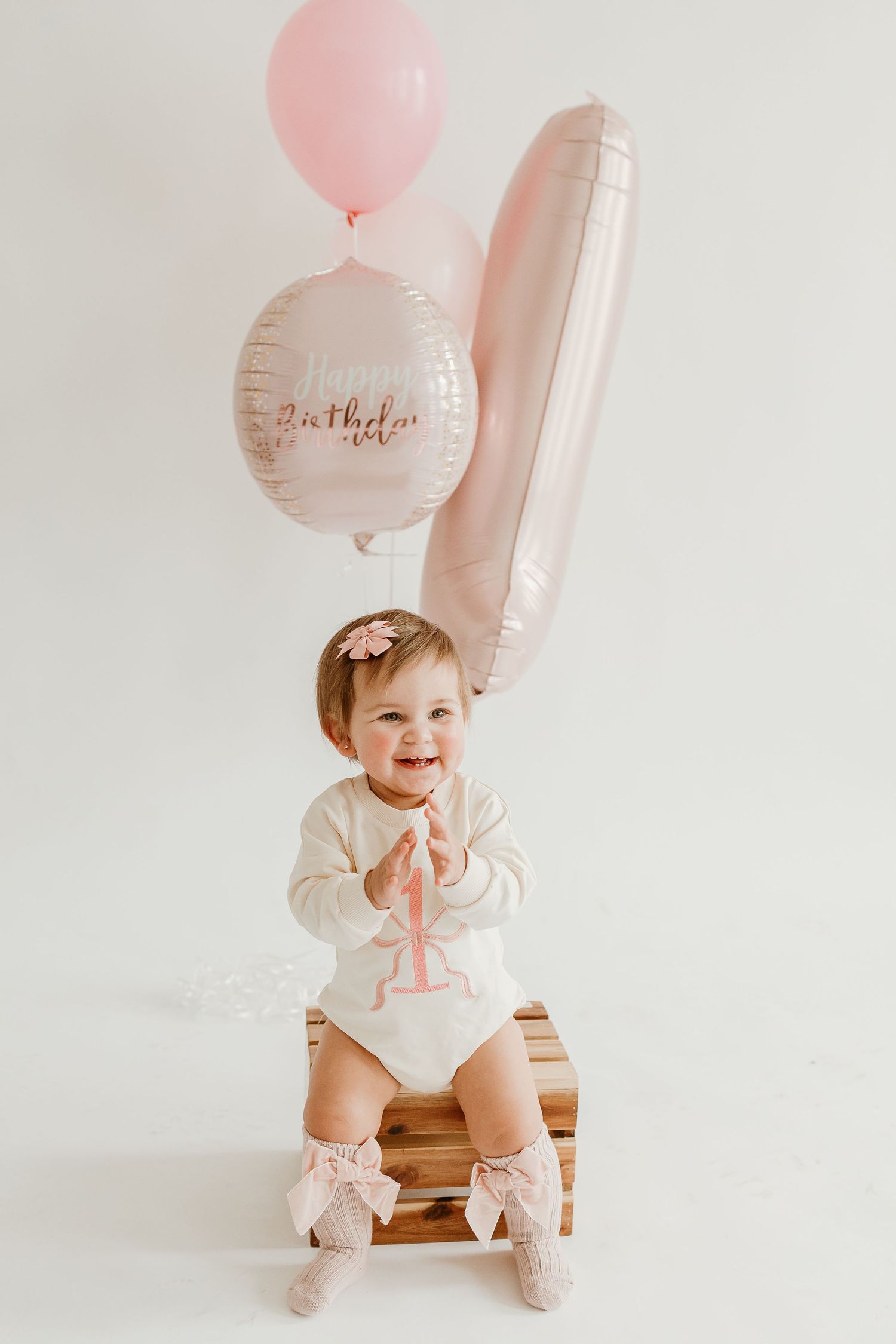 Baby in white outfit sits on wooden chair with pink birthday balloons.