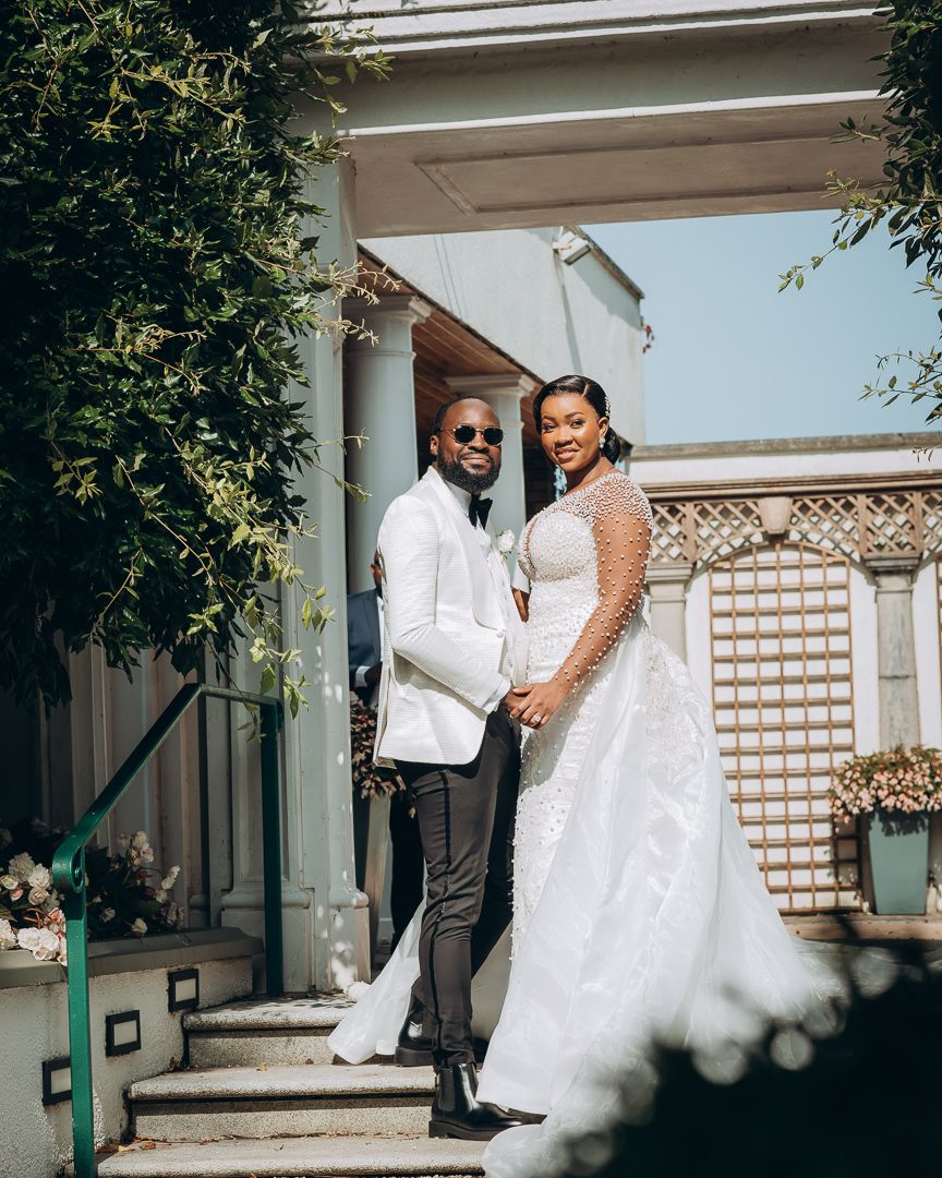 Elegant wedding couple poses on steps of venue with bride in white gown and groom in white blazer against brick wall.