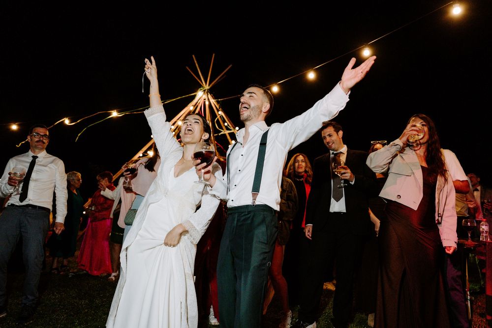 Joyful bride and groom celebrate outdoors at night with guests, string lights, and a tipi in the background.