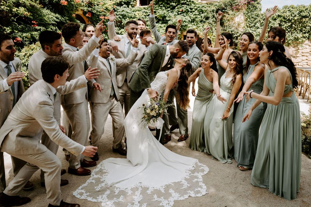 Wedding party celebrating bride and groom dip kiss, groomsmen in tan suits, bridesmaids in sage green gowns, outdoor venue.