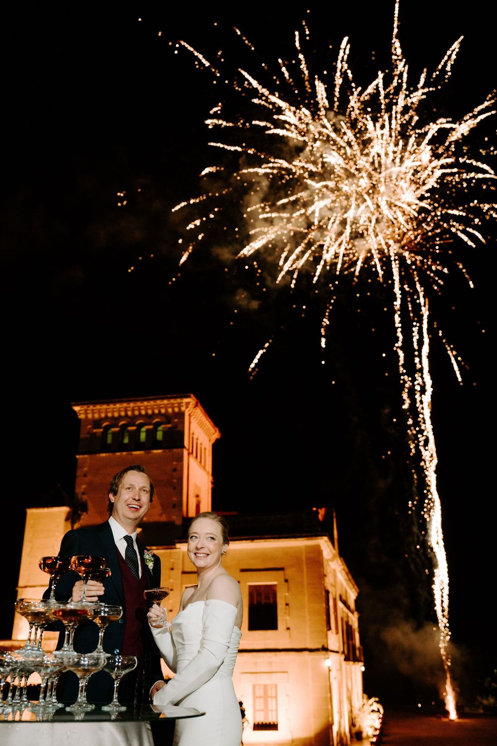 Bride and groom pose by champagne tower with fireworks bursting over illuminated castle at night.