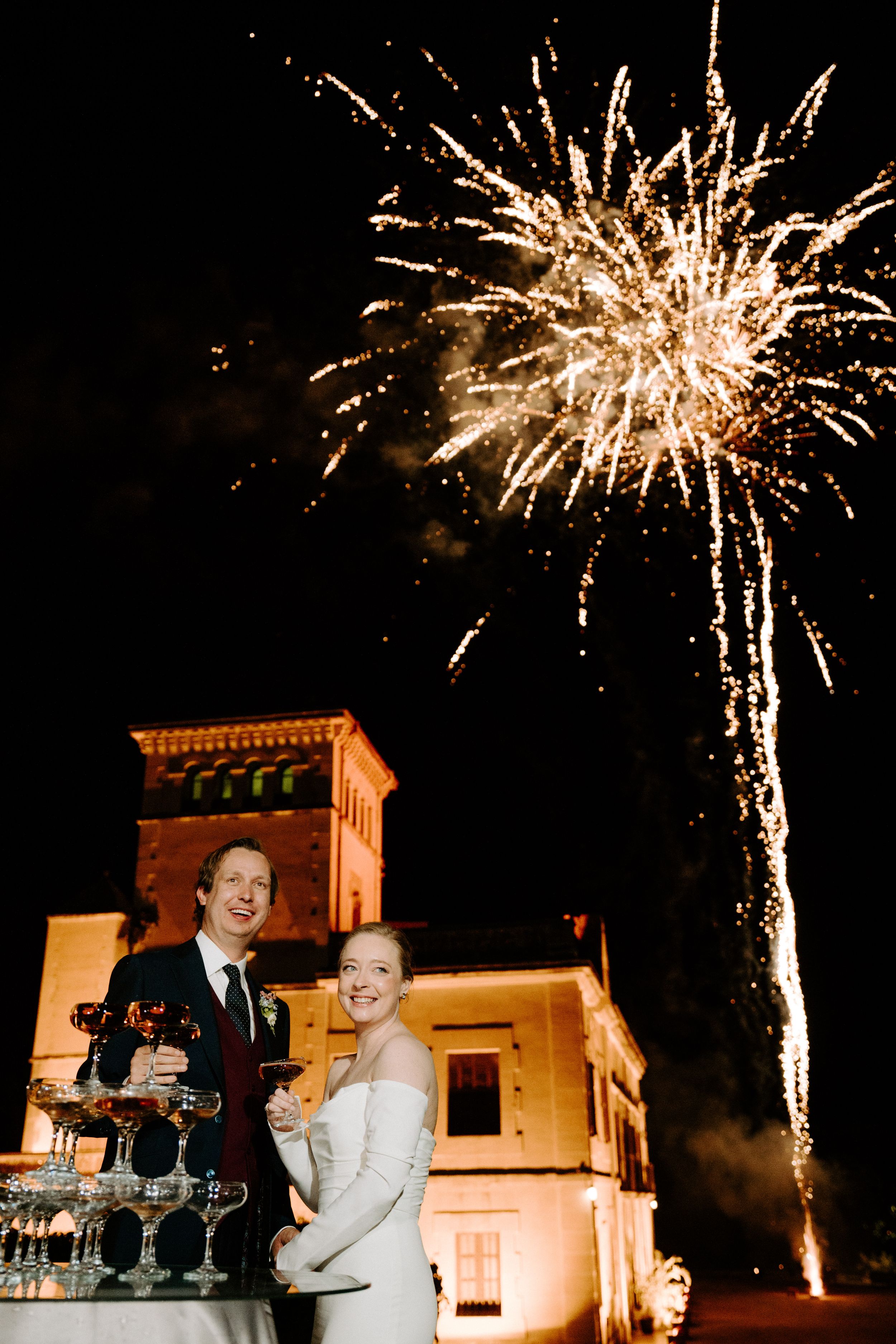 Bride and groom pose by champagne tower with fireworks bursting over illuminated castle at night.
