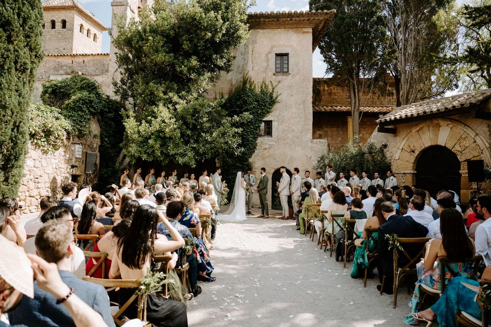 Outdoor wedding ceremony at a rustic stone villa with guests seated in wooden chairs along a sunlit aisle.