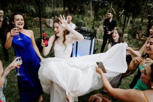 Wedding celebration with guests helping lift bride's flowing dress outdoors.