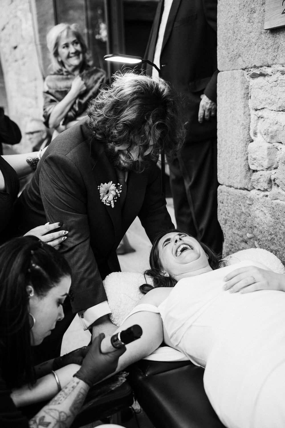 Black and white photo of a bride laughing while receiving a tattoo, surrounded by guests at a wedding.