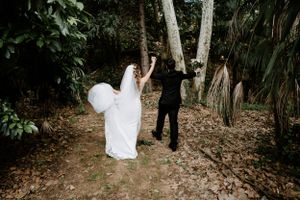 A couple poses playfully in a lush forest setting during what appears to be a wedding photoshoot.