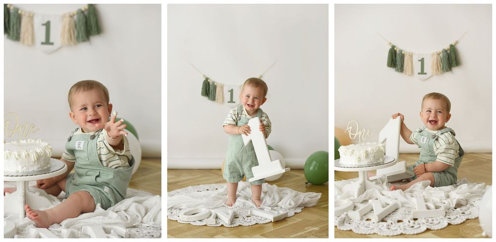 Baby in mint green overalls sits on floral blanket during first birthday photo session with cake and decorative banner.