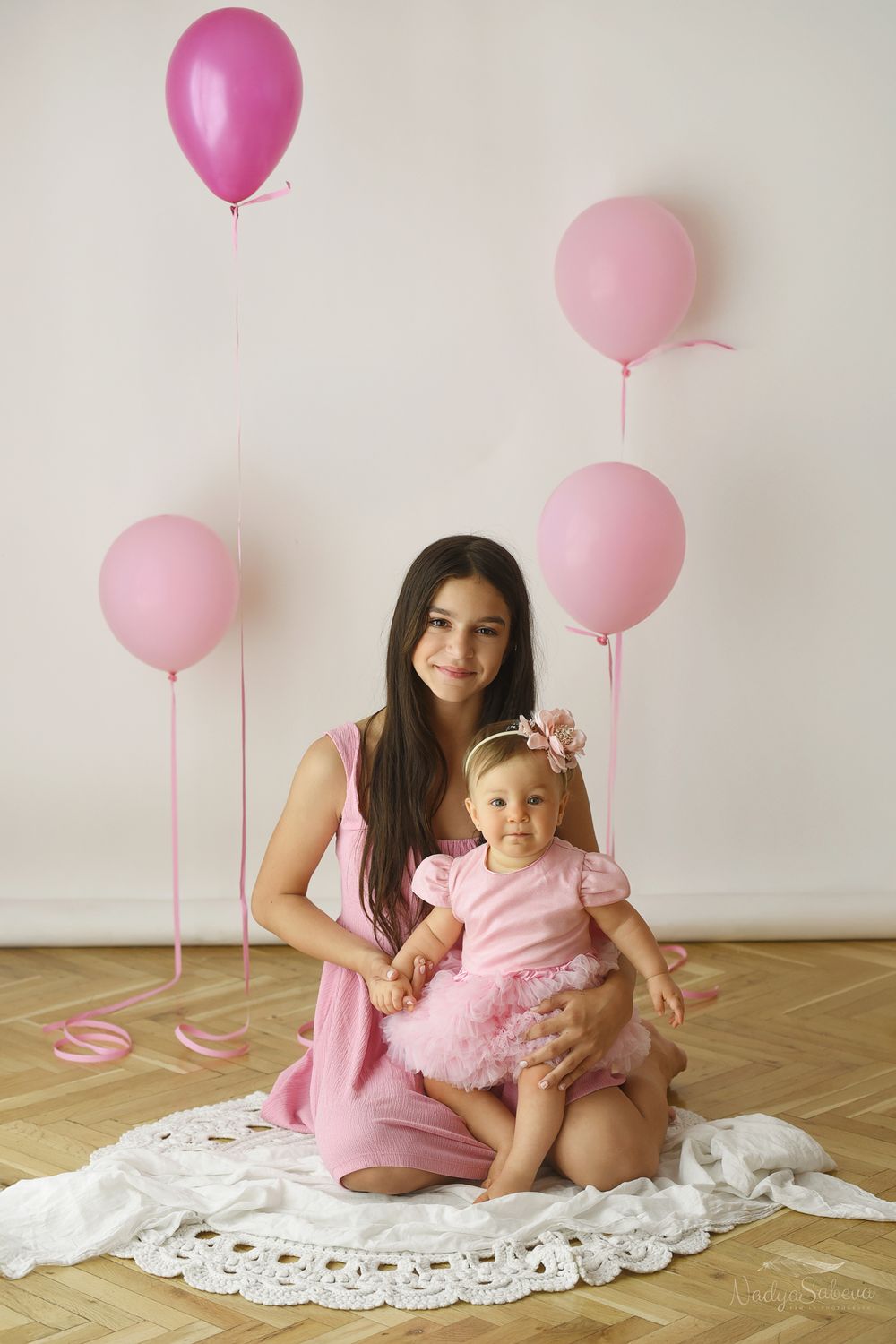 Pink balloons float above a white lace blanket in a minimalist photo studio setting with hardwood floors.