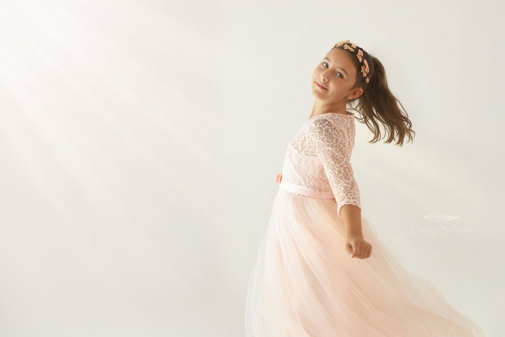 A young dancer in a flowing pink tulle dress twirls gracefully against a white studio backdrop.