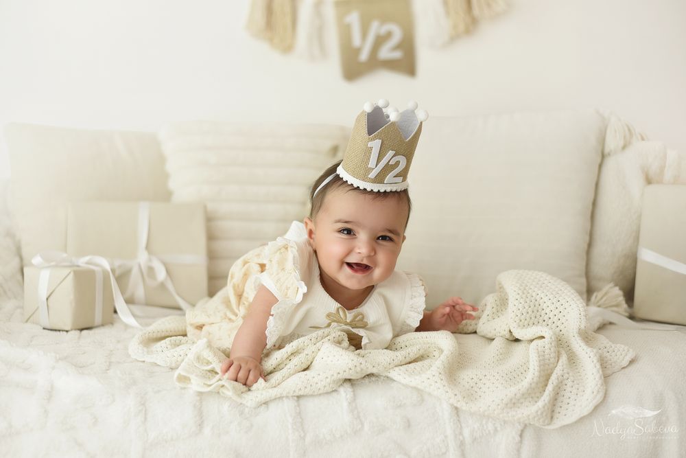 A baby in white clothing smiles while laying on cream-colored bedding in a bright, airy setting.