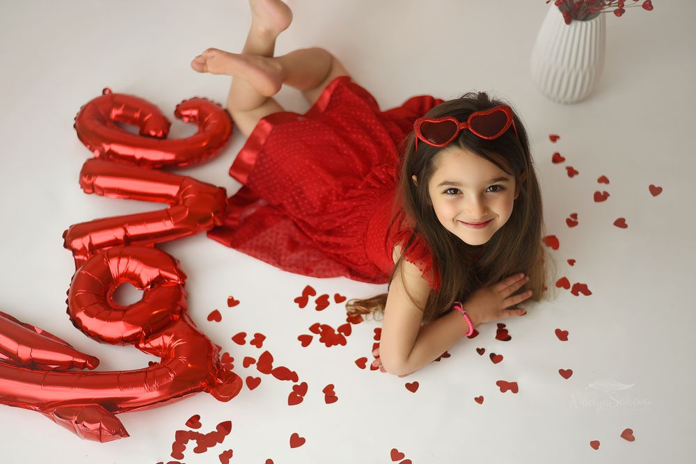 A romantic Valentine's Day themed photoshoot with scattered red rose petals and red ribbon on white fabric.