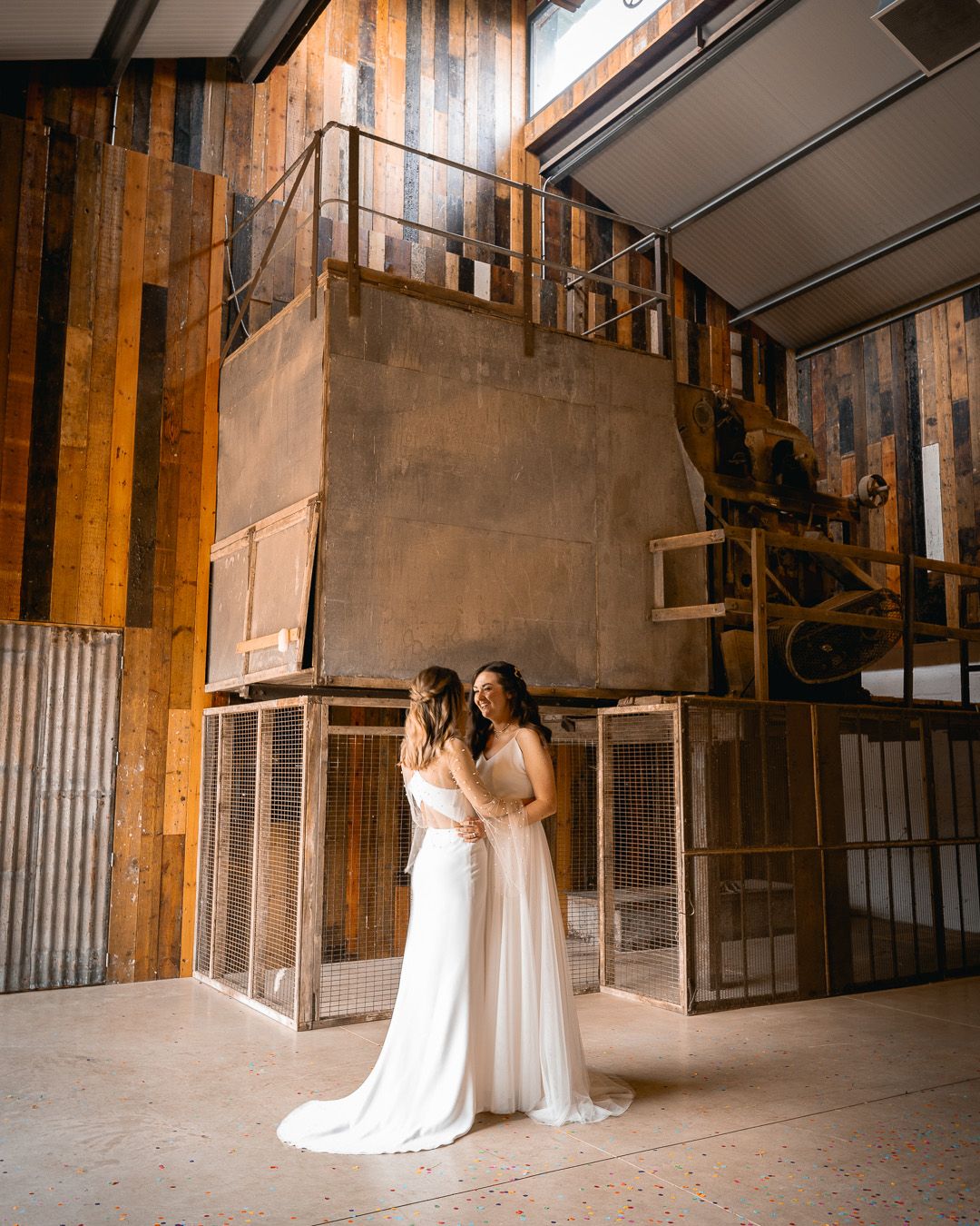 Two people in white wedding dresses stand together in an industrial-style venue with exposed brick walls.