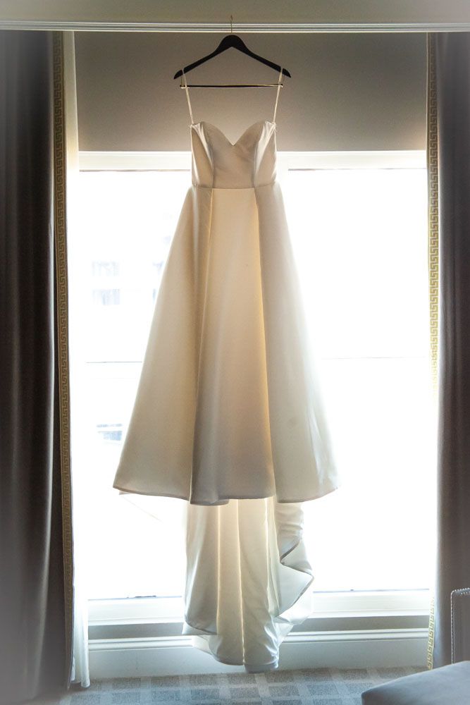 Elegant white satin A-line wedding dress with sweetheart neckline hanging on a hanger in front of a bright window.