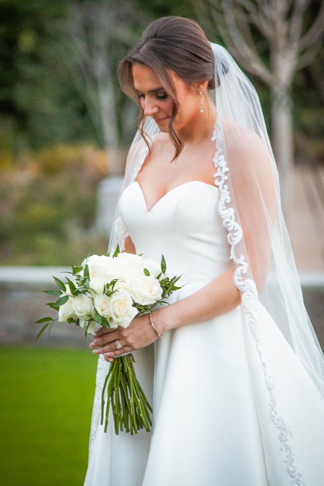 Bride in strapless white gown and lace-trimmed veil holds white rose bouquet, smiling downward outdoors.