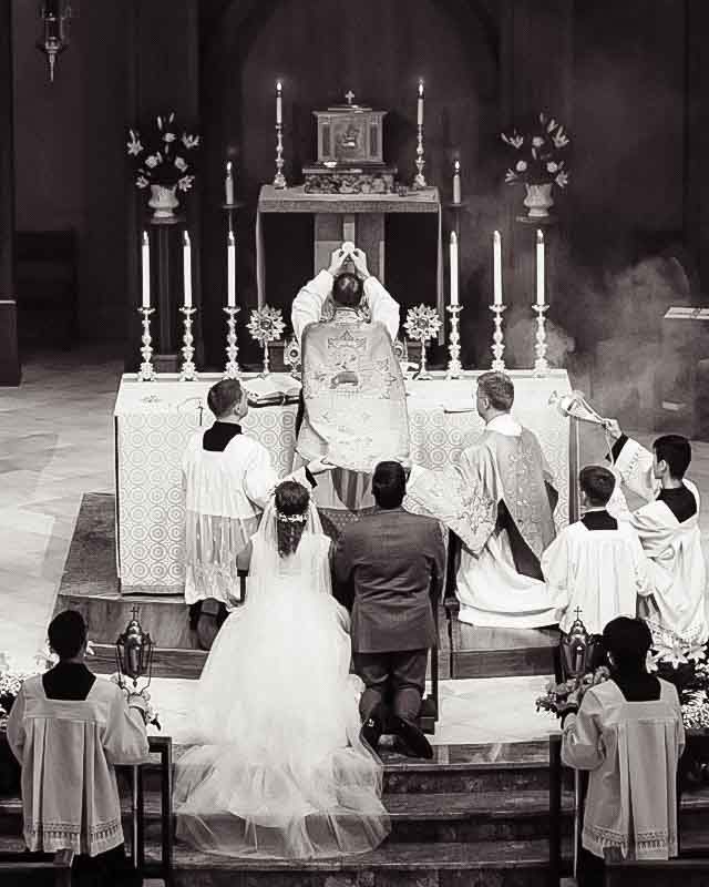 Black and white photo of a traditional Catholic wedding ceremony at the altar with priest, bride, groom, and altar servers.
