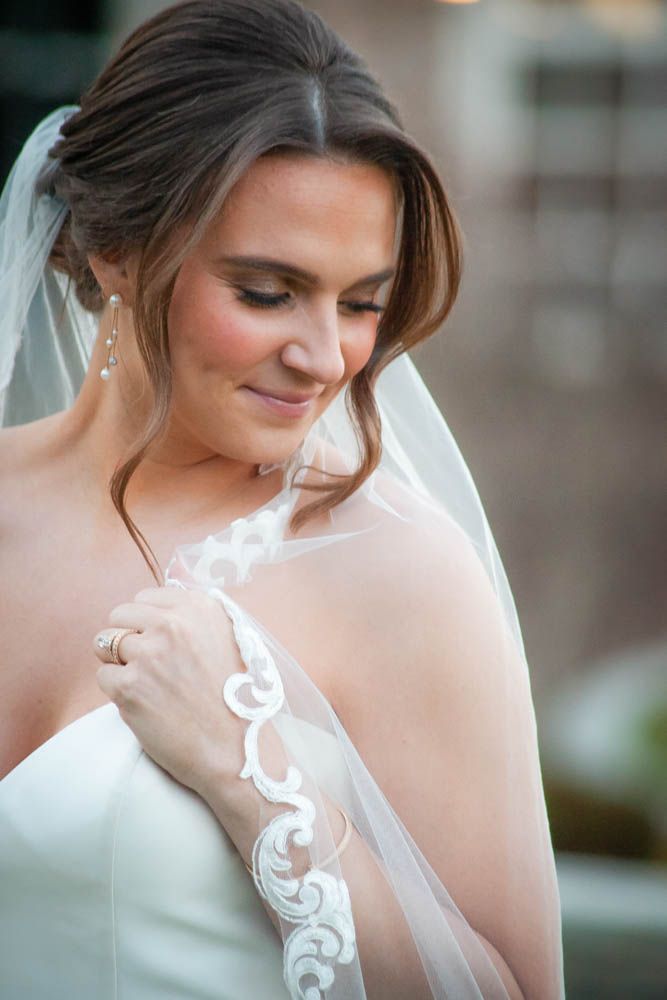 Bride in white gown and lace-trimmed veil looks down gracefully, wearing drop earrings and a diamond ring.