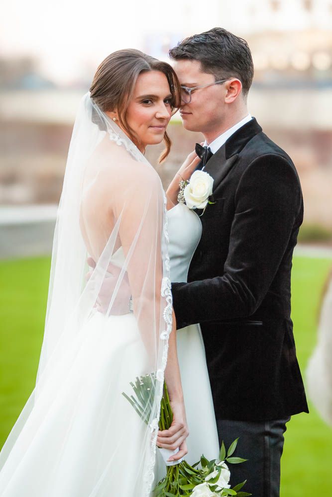 Bride and groom in black tuxedo share a romantic moment outdoors, her lace veil flowing behind them.
