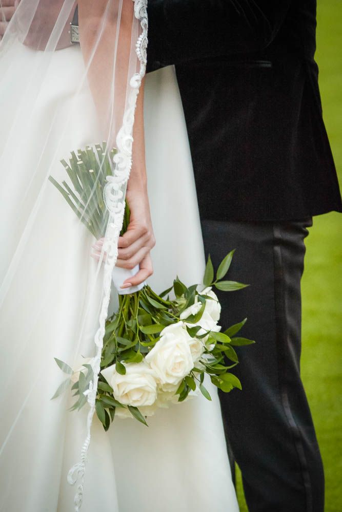 Bride in white gown holding elegant white rose bouquet with greenery beside groom in black suit at wedding ceremony.