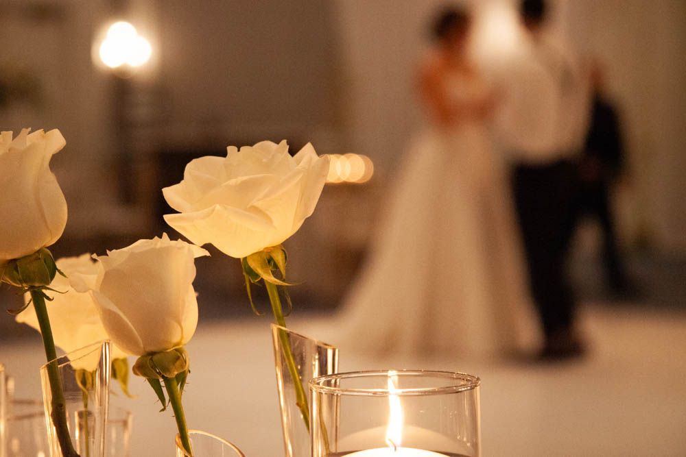 White roses in glass vases with a lit candle, couple dancing in soft focus background at a romantic wedding reception.