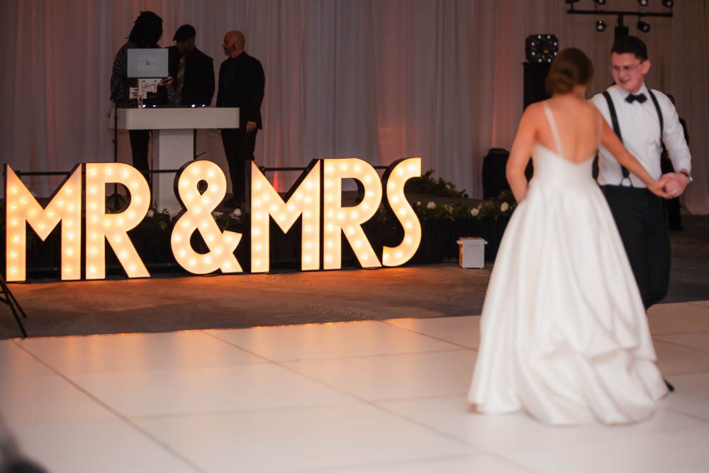 Newlyweds share first dance in front of illuminated MR & MRS marquee letters at elegant wedding reception.