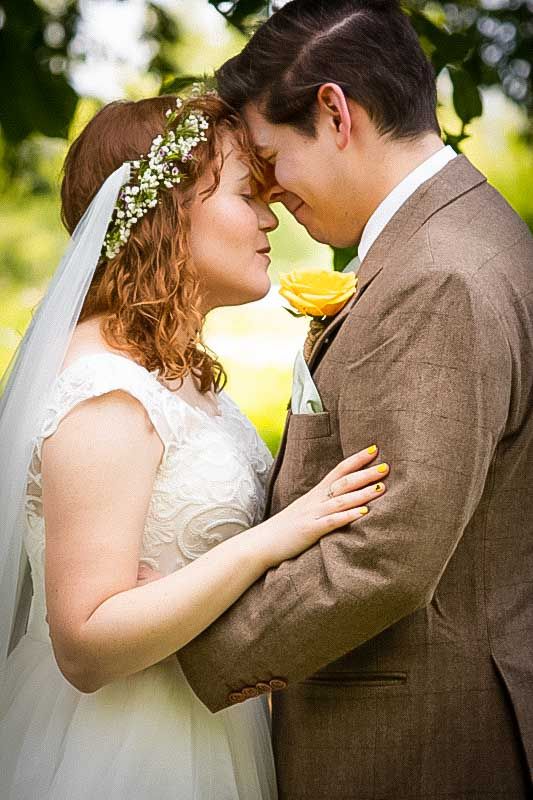 Bride and groom touching foreheads outdoors, bride wearing floral crown and veil, groom in brown suit with yellow rose boutonniere.