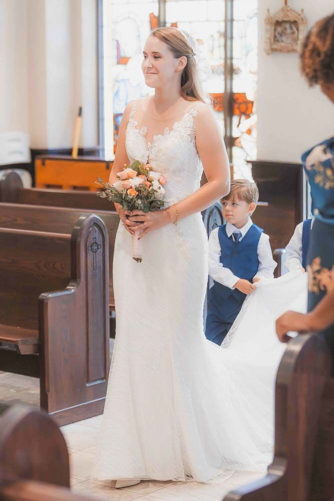 Bride in lace wedding gown holding bouquet walks down church aisle with young ring bearer in blue suit.