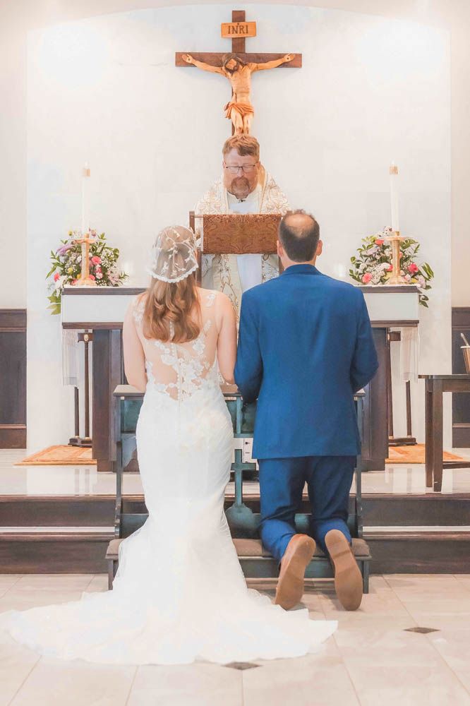 Bride and groom kneeling at church altar during Catholic wedding ceremony with priest and crucifix.