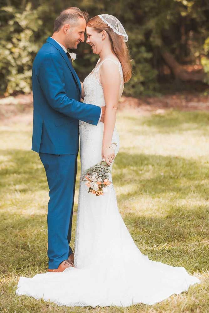 Bride and groom share a romantic moment outdoors, she in a white gown with bouquet, he in a blue suit.